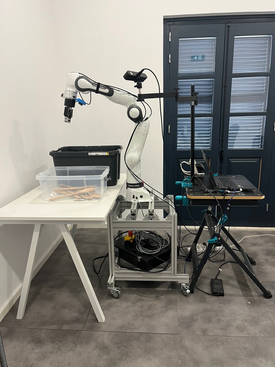 PhAIL v1.0 workcell: Franka Research 3 on aluminum-extrusion mobile base, source and destination totes on a table, separate stand with exterior camera, controller laptop, and VR teleop equipment.